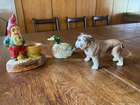 Four vintage ceramic and porcelain figurines displayed on a wooden table including gnome, duck, bulldog, and seagull