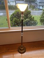 Full view of the tall antique floor lamp standing by a window, showing metal base and frosted glass bowl lampshade illuminated.