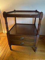 Antique mahogany side table with turned legs, two drawers with brass drop handles, and raised edge top shelf with rounded finials.