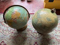 Two Soviet era globes side by side showing world maps, one with green metal stand and wooden base, the other with metal arm mount and wooden base, on patterned carpet.