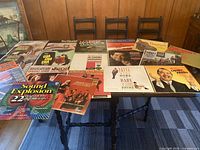 Photo showing 20 vinyl records spread over a table with visible wear on covers, artwork includes colorful graphics, title text, and photographic portraits with some album names visible.