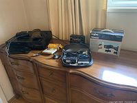 Wide shot of miscellaneous electronics on a wooden dresser including boxed Argus TeleVideo Producer, cables, camera remote, Carefree battery box, yellow packaged battery accessory, and small black/grey electronic device in a case.
