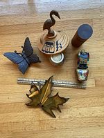 All six items displayed on a wooden surface with a ruler for scale. Items are a metal butterfly, metal maple leaf, wooden cylinder, wooden crane art piece, cow bell, and candle holder.