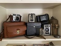 Shelf with Kodak Brownie cameras, Kodak collapsible camera, leather camera cases, Kodak film canister, and Canadian military field canteen.