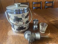 Full view of vintage metal ice bucket showing classic cars and metal handle, with metal jigger, glass shot glass, and metal strainer in front
