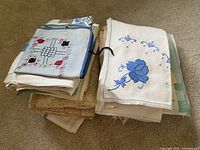 Stack of folded vintage linens including embroidered pieces with blue floral patterns, lace-bordered linens, and other assorted cloths.