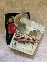 Metal tin box with painted historical guard scene on lid, partially open showing contents inside including red cigarette box and small purse.