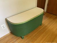 Wicker bench with closed top showing green wicker sides, white cushioned top, and rounded shape on wooden floor.