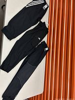 Three pairs of black Adidas men's sweatpants laid side by side showing different styles.