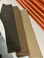 Three pairs of men's trousers in earth tones—dark olive, brown, beige—laid side by side foreground to background.