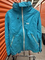Blue Helly Hansen jacket with front zipper, two zippered pockets, adjustable hood, and white HH logo on chest.