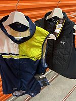 Photo of two sleeveless body warmer vests hanging on metal rack, one multicolor cycling vest by 2XU and one black Under Armour vest.