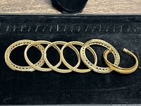 Seven gold bracelets arranged on a black velvet pad, showing details of open lattice and filigree designs on four bangles and the solid cuff shape of one bracelet