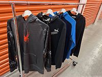 Four men's hoodies hanging on a rack: black Spider hoodie with red spider graphic, black Puma hoodie, blue and black hoodie, solid black hoodie