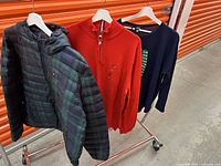 Three men's clothing items on hangers on a clothes rack: a Tommy Hilfiger plaid puffer jacket, a red quarter-zip sweater, and a navy blue long sleeve top.