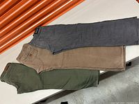 Three pairs of men’s jeans in gray, brown, and green colors laid out horizontally showing waistband and part of pant legs.