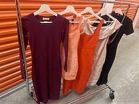 Five designer women's dresses hanging on white hangers, showing various colors and styles including burgundy, peach, orange, off-white, and black.