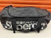 Front side of black Superdry waterproof duffel bag with white branding in Japanese and English, showing carry handles.