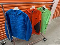 Full view of three men's Under Armour tops hanging on rack: blue insulated jacket, orange quarter-zip pullover, green long sleeve shirt.