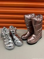 Two pairs of women's silver metallic snow boots shown side by side against an orange background, one pair mid-calf height and one pair ankle height with thick sole