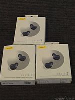 Three unopened boxes of Jabra Elite 3 earbuds arranged together on a dark carpet. Shows the front of the boxes with product images and branding.
