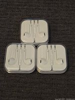 Photo of three pairs of Apple wired earbuds in original plastic square cases with transparent lids, arranged on dark fabric surface.