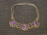 Full necklace with gold-tone metal work and three large purple cabochon stones with smaller rhinestones on a textured brown surface.