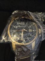 Close-up of the men's Ben & Sons watch still sealed in plastic, showing the black dial with white numerals, gold-tone case, and brown leather strap.