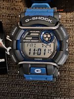 Front view of the Casio G-Shock men's digital wristwatch with a blue strap, displaying time, date, and various function labels on the dial.