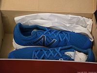 Pair of blue New Balance men's sneakers inside open box sitting on white tissue paper. Each shoe has blue mesh upper, white midsole, blue laces, and large reflective 'N' logo on side.