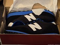 Pair of brand new black New Balance men's sneakers with blue midsoles and white logos inside original box with tissue paper.