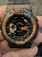 Close-up of the black Casio G-Shock watch with orange accents and analog-digital dial held in hand.