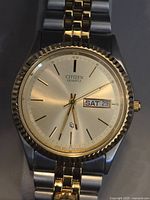 Front view of Citizen men's quartz watch showing dial, gold-tone bezel, and day/date display at 3 o'clock.