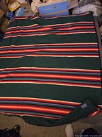 Full view of large Pendleton blanket spread out, showing dark green base with horizontal red, orange, white, and black stripes.