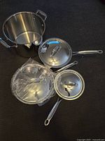 Four-piece stainless steel cookware set including saucepan with lid, frying pan with lid, stock pot, and smaller pan, all on dark surface with plastic wrap on some parts.