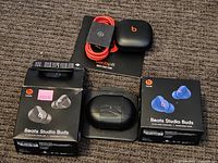 Photo showing two Beats Studio Buds retail boxes, one with black earbuds and one with blue earbuds, one charging case out of box, Beats Pill Quick Start Guide booklet, and red charging cable.