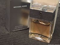 Michael Kors perfume bottle with gold-colored liquid inside and transparent cuboid cap, alongside its original black box with white text.
