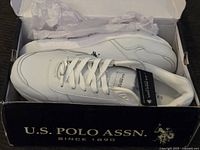 Top view of new white U.S. Polo Assn. sneaker inside partially open black shoebox with brand name on front
