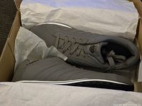 Pair of grey K-Swiss sneakers sitting in a shoebox with tissue paper inside, shoes appear new and unworn.