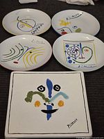 Five decorative plates laid out showing colorful Picasso-inspired abstract designs and signatures