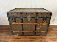 Front view of antique trunk showing two large metal locking clasps and keyhole lock center. Three wood slats horizontally across front, black metal edging and rivets.