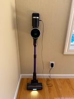 Cordless stick vacuum cleaner mounted on wall charging dock with LED light on brush head illuminating wooden floor.