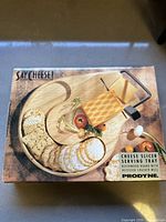 Box showing Prodyne cheese slicer serving tray with beechwood board and recessed cracker well, sealed and new in box.