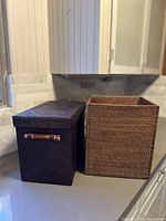 Two rectangular wicker baskets placed side by side on a kitchen counter, showing the dark brown basket with lid and handles and natural open basket.