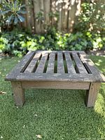 Front angle view of a small rectangular teak wood table with horizontal slats on the top surface, standing on a grassy lawn outdoors with some ivy and shrubbery in the background.