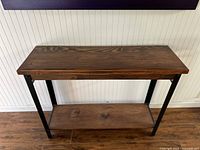 Front view of a rectangular 2-tier wooden side table with black metal legs on wood flooring against a beadboard wall.