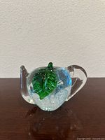 Front-side view of the clear glass teapot paperweight shaped like an apple with a prominent green leaf detail and small internal bubbles.