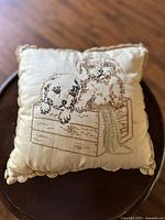 Front view of small throw pillow with embroidery of two puppies in a wooden crate, cream fabric with scalloped trim.