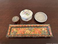 Top-down view of four items on a wooden surface: rectangular floral tray, small round trinket box, white ceramic jewelry box with 3D flowers, and a small shallow bowl.
