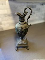 Full view of the vintage ewer urn showing its overall shape, handle, and ornate sculptural details on a countertop.